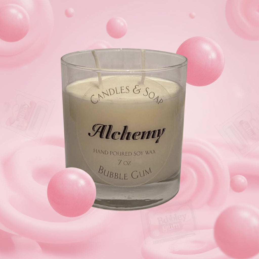 A clear glass soy wax candle with 'Alchemy' branding, labeled as 'Bubble Gum', placed on a white surface with a green plant in the background.